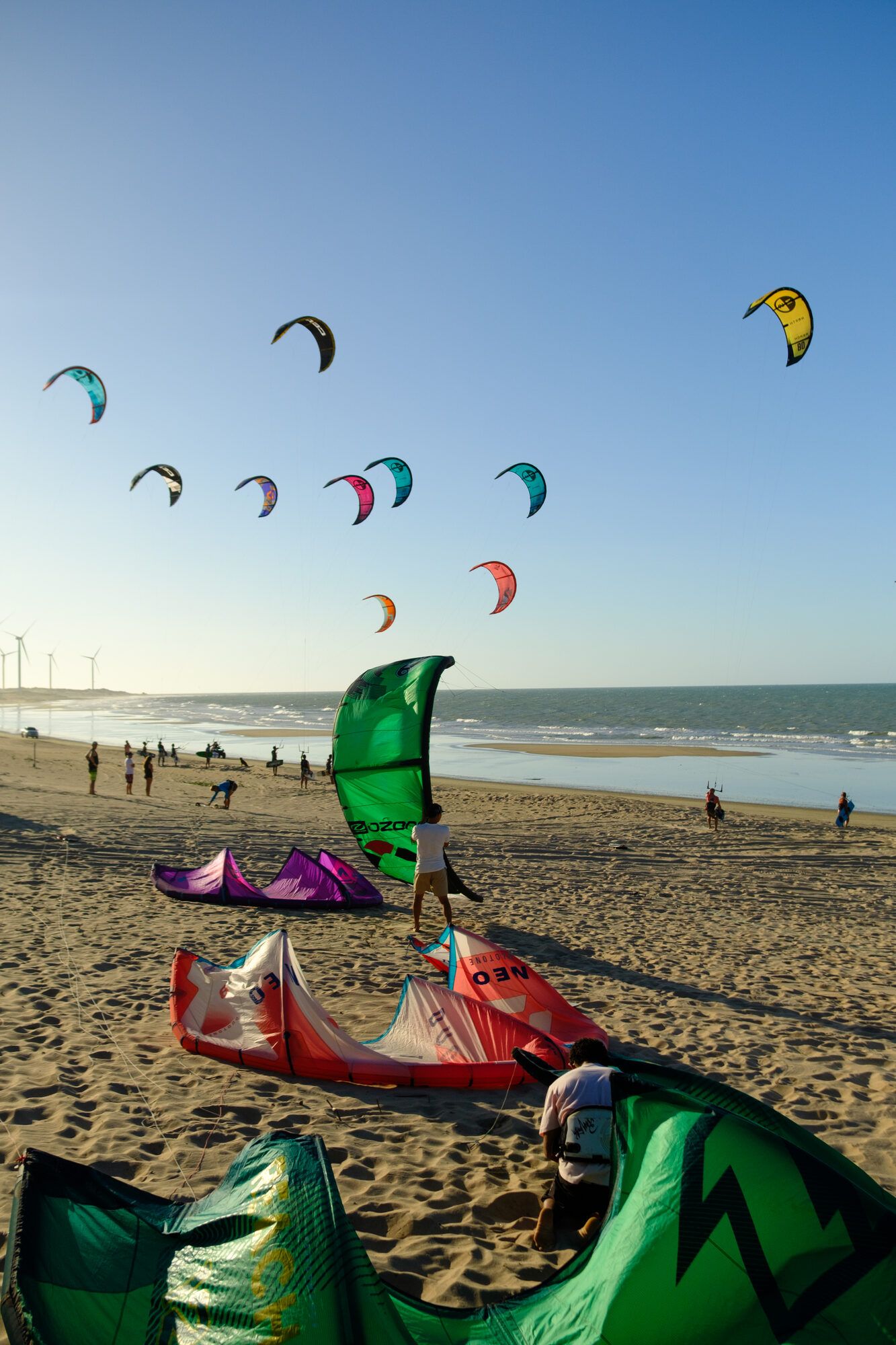 Des personnes montent leurs kites sur le sable, un parc éolien en arrière-plan.