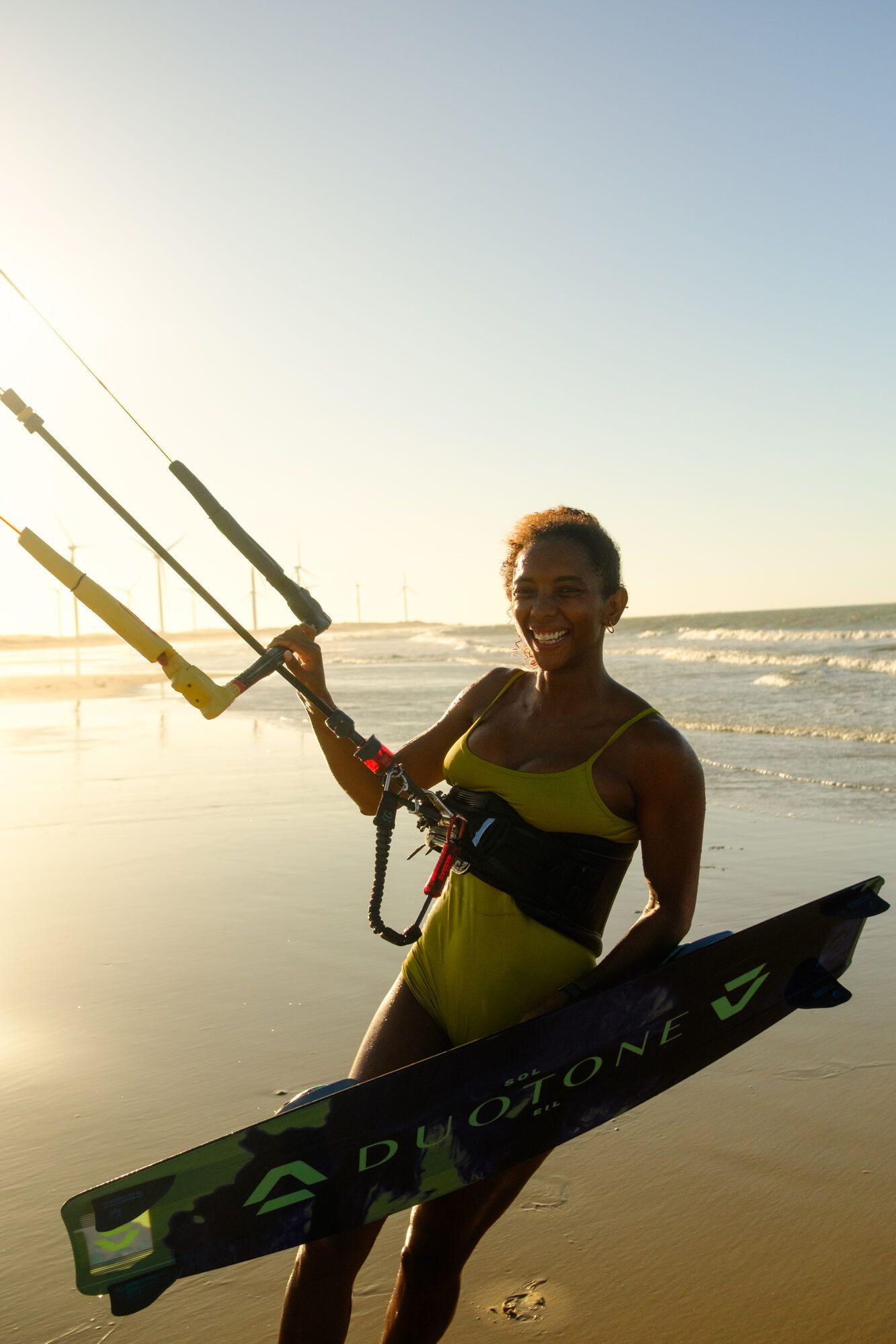Mulher sorri segurando a barra e a prancha de kite, luz dourada do fim de tarde.