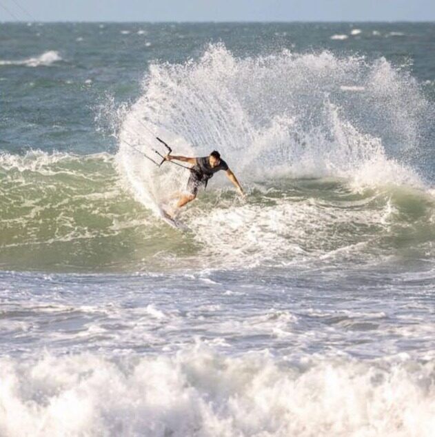 Un kitesurfeur manœuvre sur la crête d'une vague, projetant une grande gerbe d'eau.