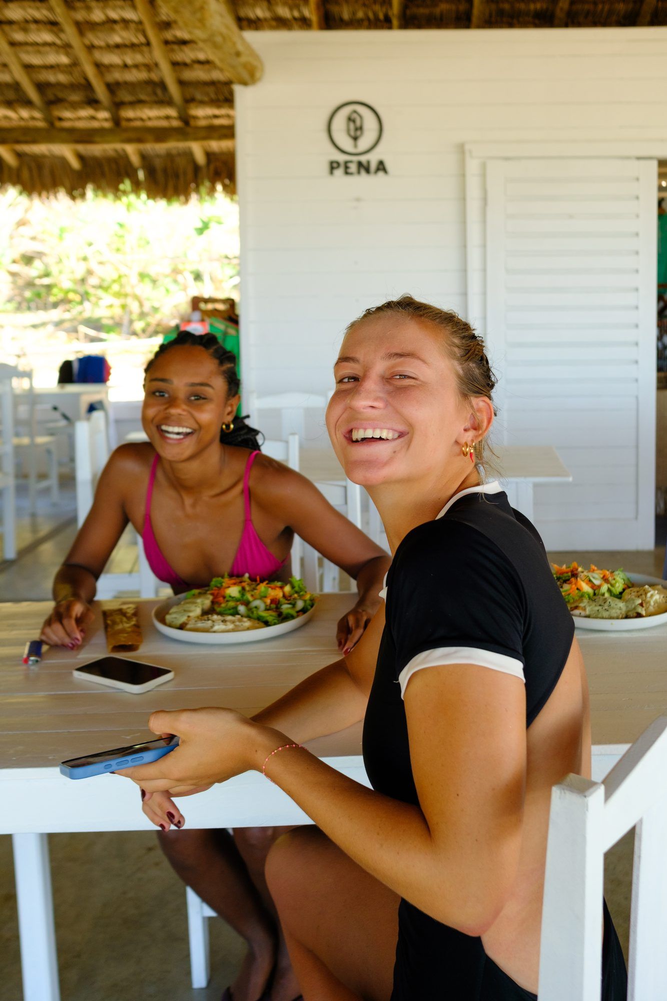 Duas amigas riem à mesa do restaurante, com pratos de salada à frente.