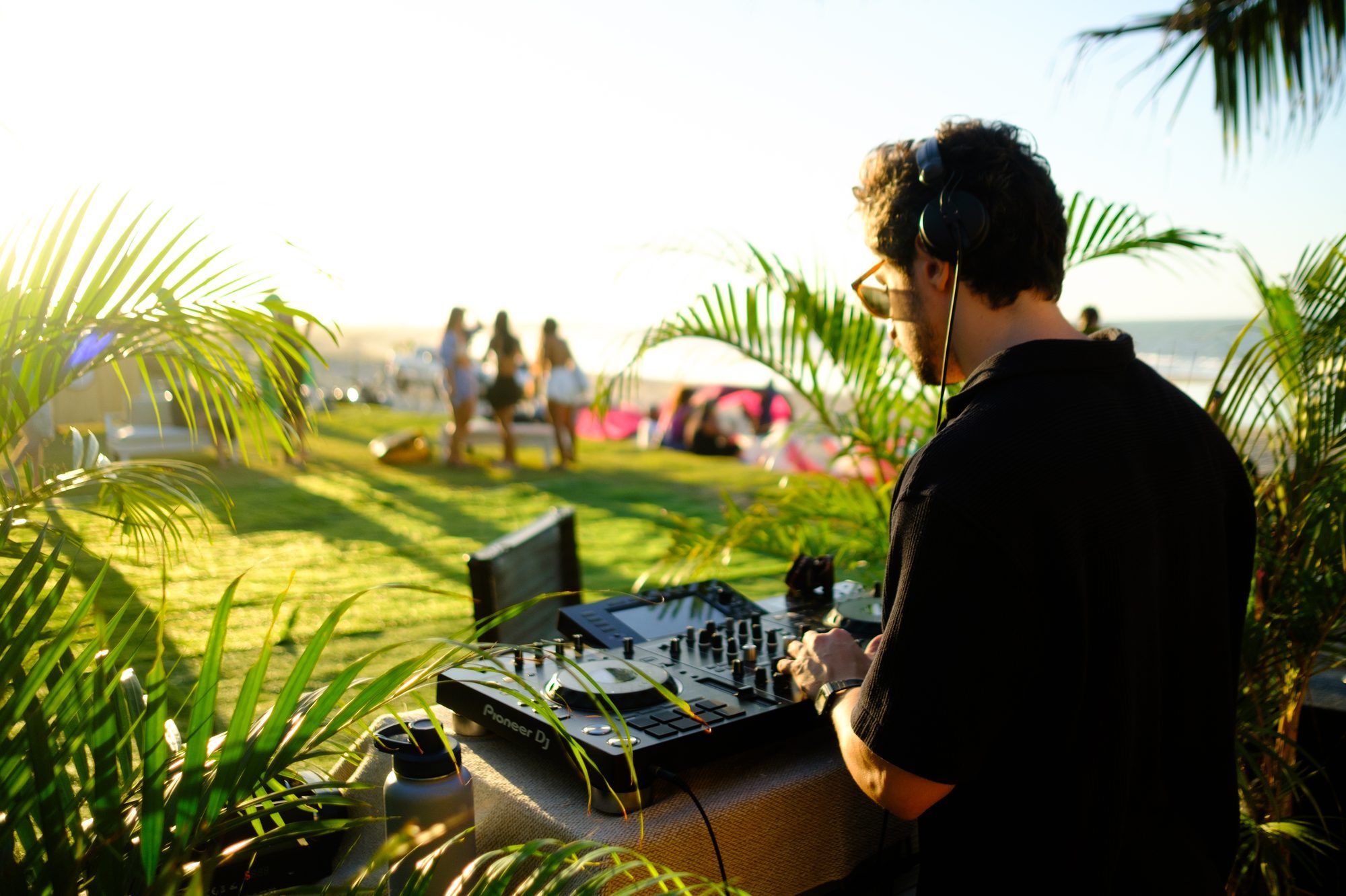 Un DJ toca entre el follaje, gente reunida en el césped al atardecer.