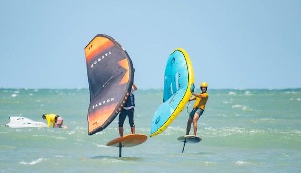 Duas pessoas praticam wingfoil lado a lado, voando sobre a água.