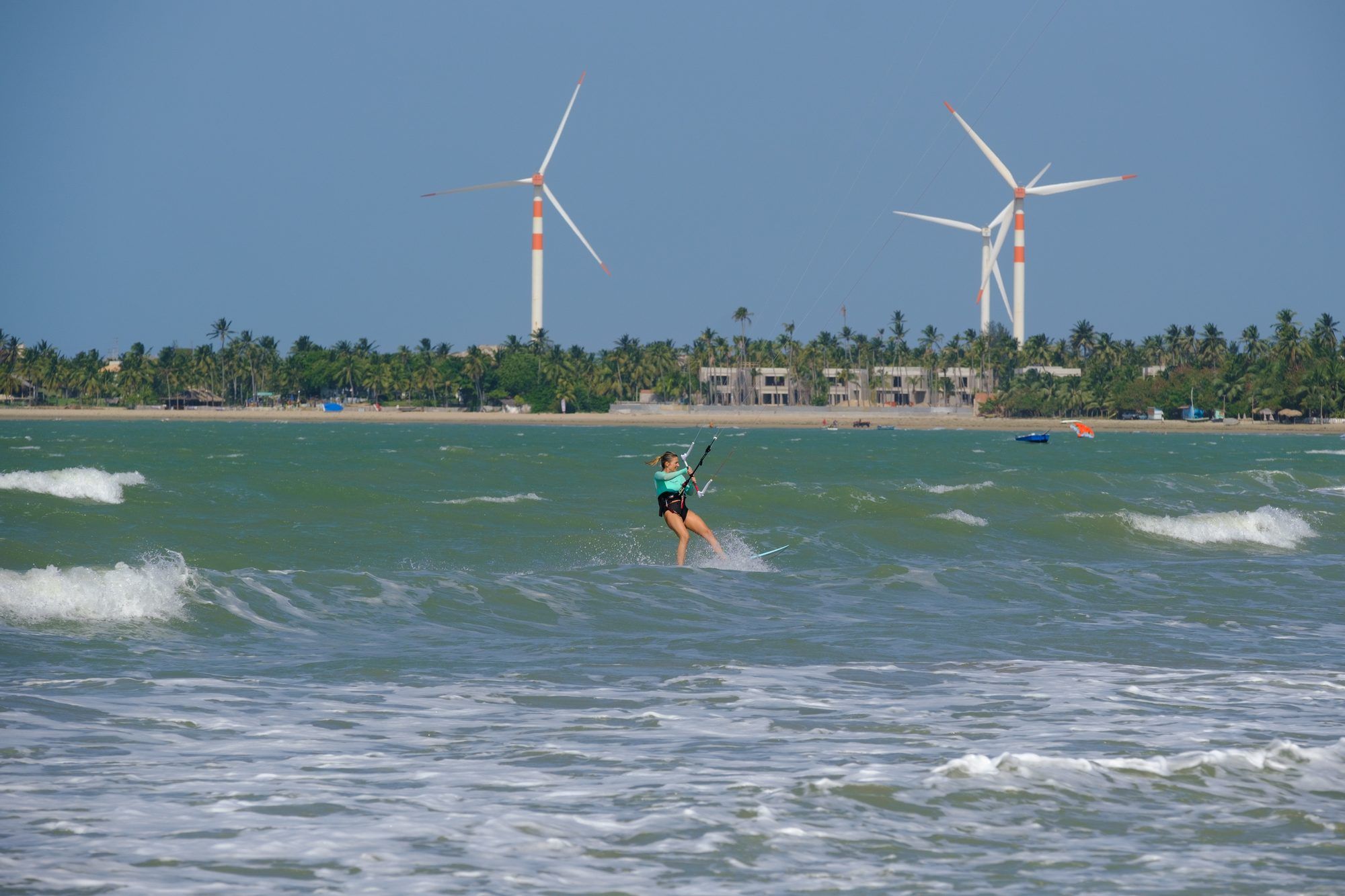 Una mujer practica kitesurf en el mar, aerogeneradores y palmeras de la costa al fondo.