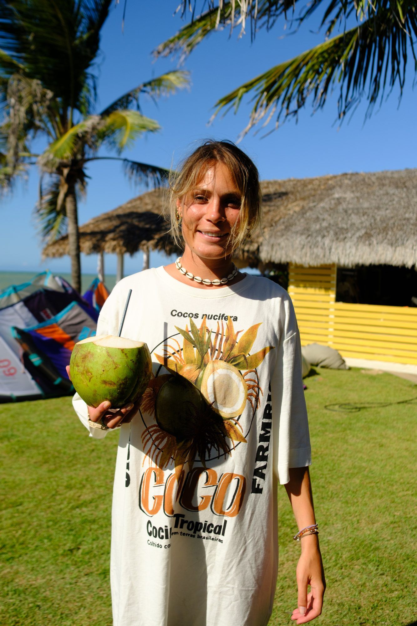 Mulher sorri segurando um coco verde, cabanas de palha ao fundo.