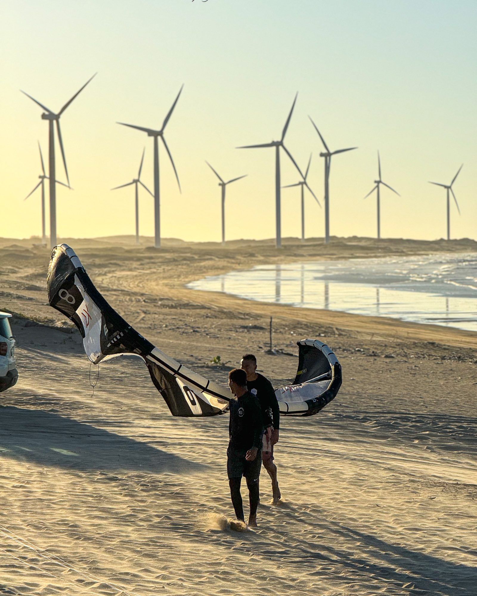 Duas pessoas caminham na praia carregando uma pipa, turbinas eólicas no horizonte ao entardecer.