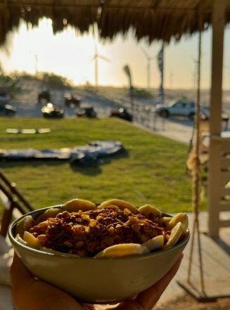 Tigela de açaí coberta de granola e banana, vista da praia ao fundo.