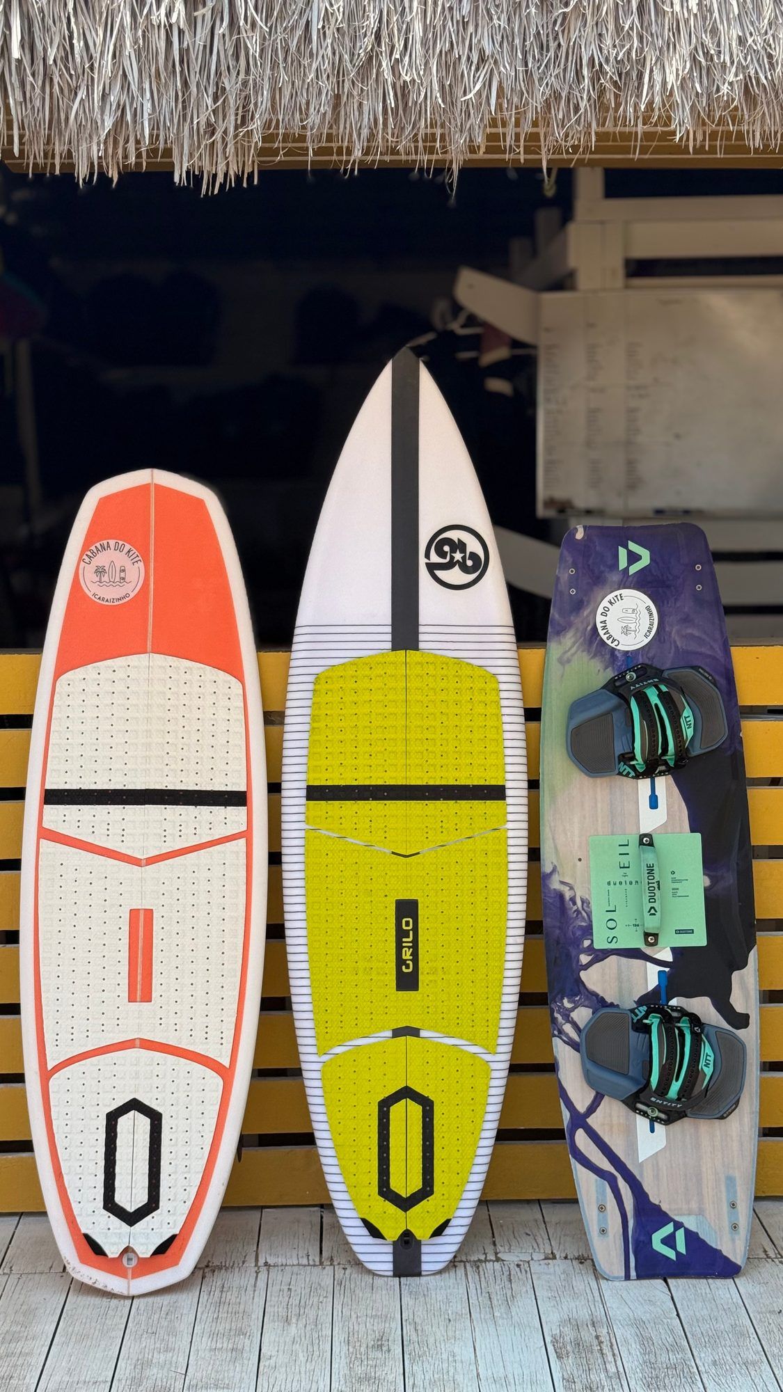 Três pranchas apoiadas lado a lado — kitesurf, surf com foil e wing.