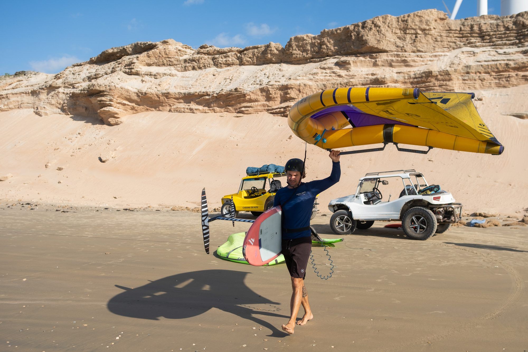 Praticante carrega uma asa de wing e uma prancha com foil pela praia, com dunas ao fundo.
